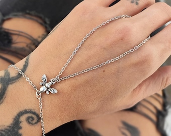 May include: A silver-toned hand chain featuring a small bee charm. The jewelry consists of a delicate link chain that wraps around the wrist and extends across the back of the hand, connecting to a central bee-shaped metal ornament.