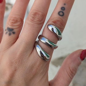 Op de afbeelding: Een zilverkleurige ring met een glad, gebogen ontwerp wikkelt zich om een vinger. De ring heeft een gepolijst, reflecterend oppervlak. De hand heeft ook een pootafdruk-tattoo en rode nagellak met zwarte stippen.