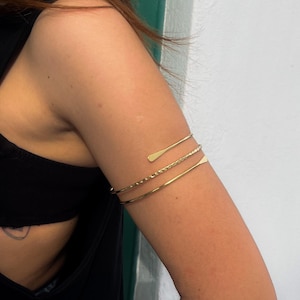 May include: Gold-toned arm cuff jewelry. The image shows three separate arm cuffs, each with a unique design. The top cuff has a flat, elongated shape, the middle cuff has a twisted rope design, and the bottom cuff is a simple band.