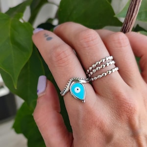 May include: A silver ring with a teardrop-shaped turquoise enamel design featuring a white and black eye. The ring is worn with two other silver rings with a beaded design. Green leaves are in the background.