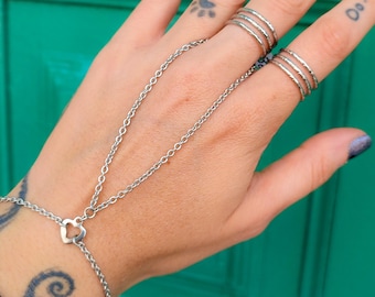 May include: A silver-toned hand chain bracelet featuring a small heart-shaped connector, connected to two sets of three stacked silver-toned rings worn on the middle and ring fingers, all crafted with a textured, metallic finish.