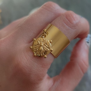 May include: Gold ring with a bee charm. The ring is wide and has a smooth, flat surface. The bee charm is detailed and hangs from the ring.