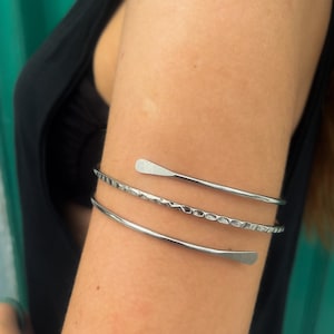 May include: Three silver arm cuffs. The top cuff has a flat, paddle-like design. The middle cuff has a textured, hammered appearance. The bottom cuff is smooth. The arm cuffs are worn on the upper arm.