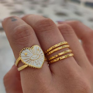 Puede incluir: Anillos dorados. Un anillo presenta un diseño blanco en forma de corazón con un ojo y la palabra "LOVE". El otro anillo tiene un diseño de banda texturizada. Joyas.