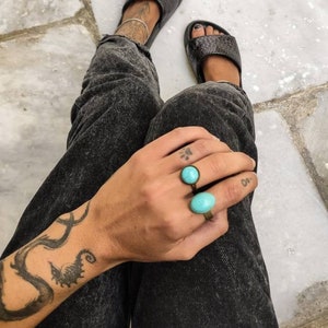 Puede incluir: Sandalias negras de cuero con un patrón texturizado. Se ven dos anillos turquesas y unos vaqueros negros. Un tatuaje es visible en el brazo.