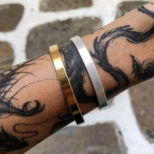 Pode incluir: S&atilde;o exibidas duas pulseiras minimalistas, uma dourada e outra prateada. A pulseira dourada est&aacute; &agrave; esquerda e a prateada &agrave; direita. As pulseiras s&atilde;o usadas em um bra&ccedil;o com uma grande tatuagem preta. O fundo &eacute; uma superf&iacute;cie de pedra branca e cinza.
