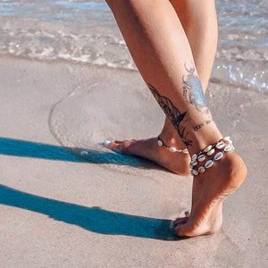 Könnte beinhalten: Fußkettchen aus weißen Muscheln werden an den Knöcheln getragen. Die Person steht an einem Sandstrand mit dem Meer im Hintergrund. Ein Tattoo ist am Knöchel zu sehen. Das Bild ist bei natürlichem Licht aufgenommen.