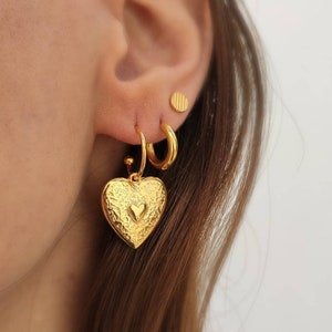 Puede incluir: Un conjunto de pendientes dorados. Un pendiente presenta un colgante en forma de coraz&oacute;n con un dise&ntilde;o en relieve y un coraz&oacute;n m&aacute;s peque&ntilde;o en el centro. Los otros pendientes son un aro peque&ntilde;o y un peque&ntilde;o perno redondo. Los pendientes se llevan en una oreja.