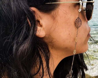 May include: A gold-toned metal eyeglass chain featuring delicate leaf-shaped charms and small circular links. The chain attaches to the temples of gold-rimmed sunglasses with white rubber loops, hanging down along the side of the face.