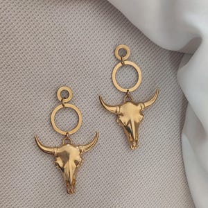 K&ouml;nnte beinhalten: Ein Paar goldfarbene Ohrringe mit einem Bullensch&auml;del-Design. Jeder Ohrring hat einen kreisf&ouml;rmigen oberen Teil, der mit einem Bullensch&auml;del-Anh&auml;nger mit gebogenen H&ouml;rnern verbunden ist.