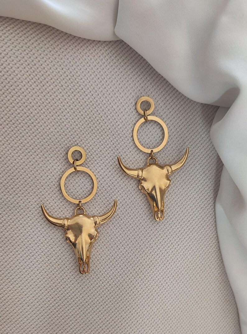 K&ouml;nnte beinhalten: Ein Paar goldfarbene Ohrringe mit einem Bullensch&auml;del-Design. Jeder Ohrring hat einen kreisf&ouml;rmigen oberen Teil, der mit einem Bullensch&auml;del-Anh&auml;nger mit gebogenen H&ouml;rnern verbunden ist.