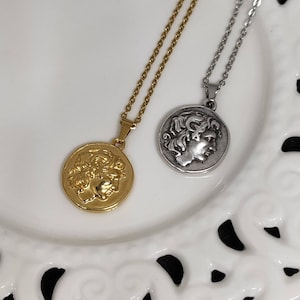 Puede incluir: Dos collares con colgantes de monedas. Uno es dorado, el otro plateado. Cada colgante presenta el perfil de una persona con cabello rizado. Los collares est&aacute;n sobre un plato blanco decorativo.