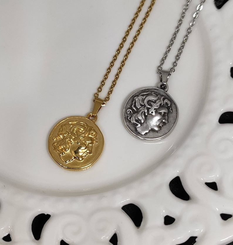 Puede incluir: Dos collares con colgantes de monedas. Uno es dorado, el otro plateado. Cada colgante presenta el perfil de una persona con cabello rizado. Los collares est&aacute;n sobre un plato blanco decorativo.