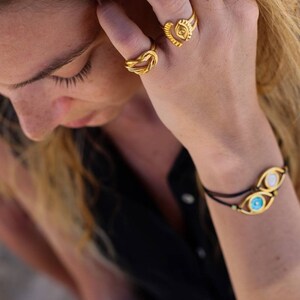 May include: A close-up shot of a person's hand adorned with gold jewelry. The hand features two rings, one a knot design and the other with a square center. A bracelet with two eye-shaped charms completes the look. The jewelry is set against a black top.