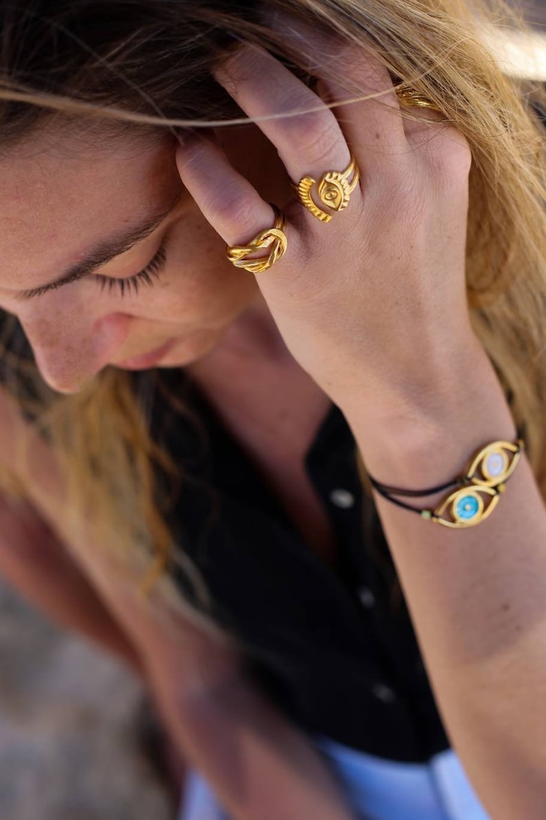 May include: A close-up shot of a person's hand adorned with gold jewelry. The hand features two rings, one a knot design and the other with a square center. A bracelet with two eye-shaped charms completes the look. The jewelry is set against a black top.