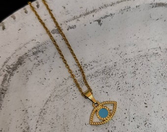 Puede incluir: Collar dorado con un colgante en forma de ojo con un centro azul claro. El colgante tiene un diseño detallado y texturizado. La cadena es delicada y fina. El collar se muestra sobre una superficie texturizada blanquecina.