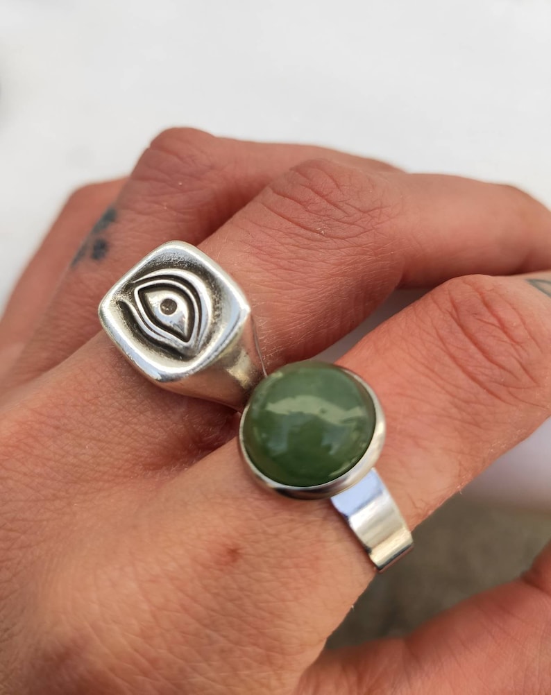 Pu&ograve; includere: Due anelli d'argento su un dito. L'anello sull'indice ha una faccia quadrata con un disegno di occhio. L'anello sul medio ha una pietra verde rotonda.