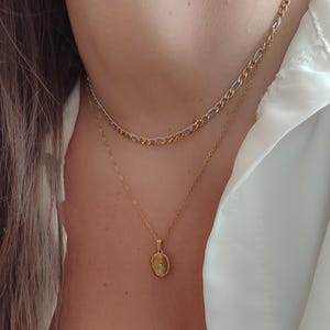 May include: A close-up of two gold necklaces. The top necklace is a chain with alternating gold and silver links. The bottom necklace has a delicate gold chain with a small, oval pendant. The necklaces are worn over a white shirt.