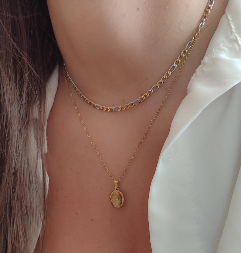 May include: A close-up of two gold necklaces. The top necklace is a chain with alternating gold and silver links. The bottom necklace has a delicate gold chain with a small, oval pendant. The necklaces are worn over a white shirt.