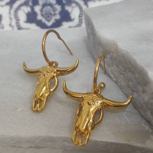 May include: A pair of gold-colored earrings featuring a bull skull design. The earrings have a hoop style with a detailed skull pendant. The earrings are displayed on a white surface with a blue and white patterned background.