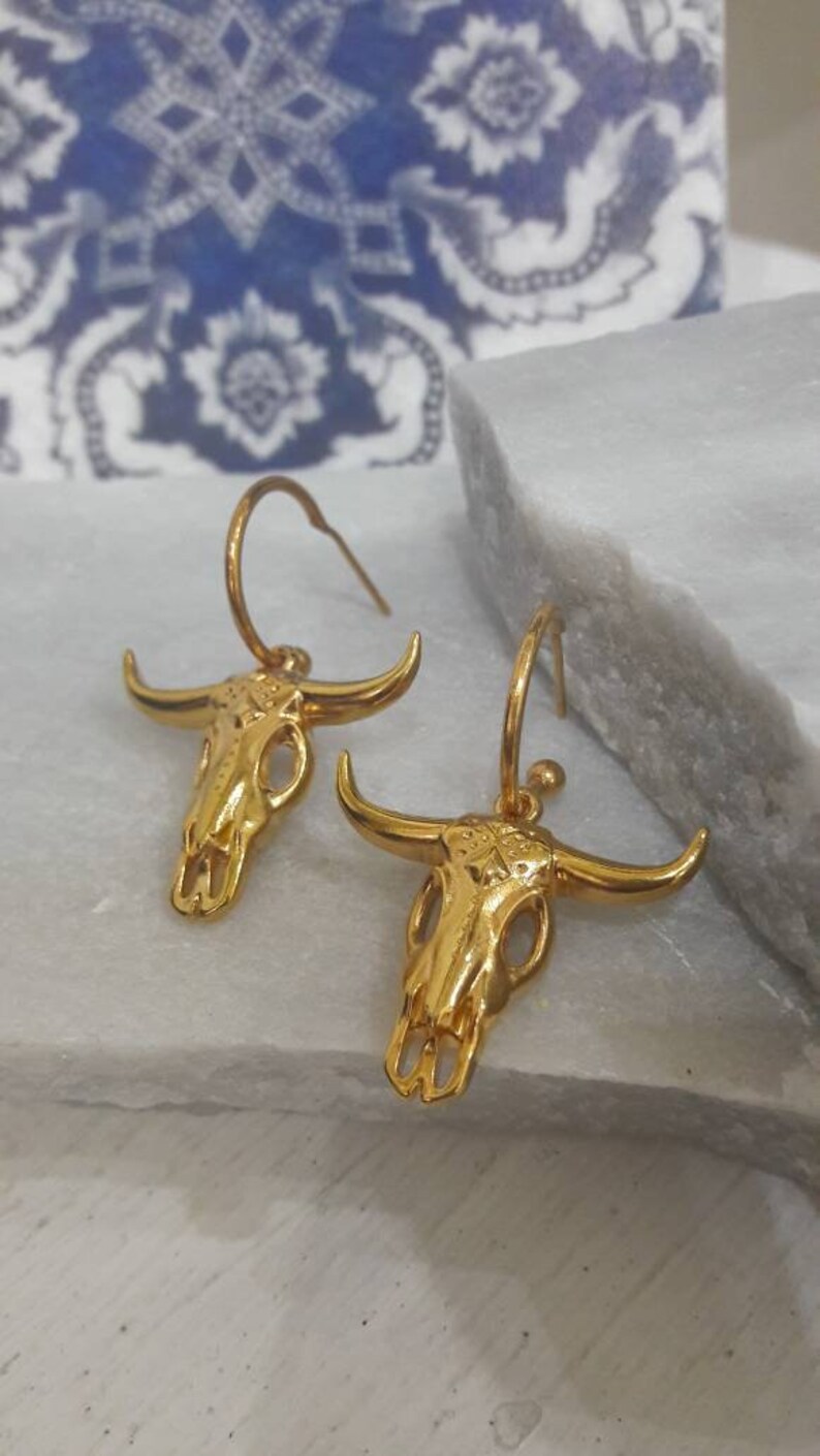 May include: A pair of gold-colored earrings featuring a bull skull design. The earrings have a hoop style with a detailed skull pendant. The earrings are displayed on a white surface with a blue and white patterned background.
