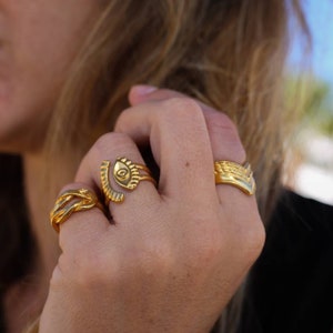 May include: A hand wearing several gold rings. The rings feature various designs, including an eye, woven patterns, and a textured band. The rings are made of a shiny gold material.