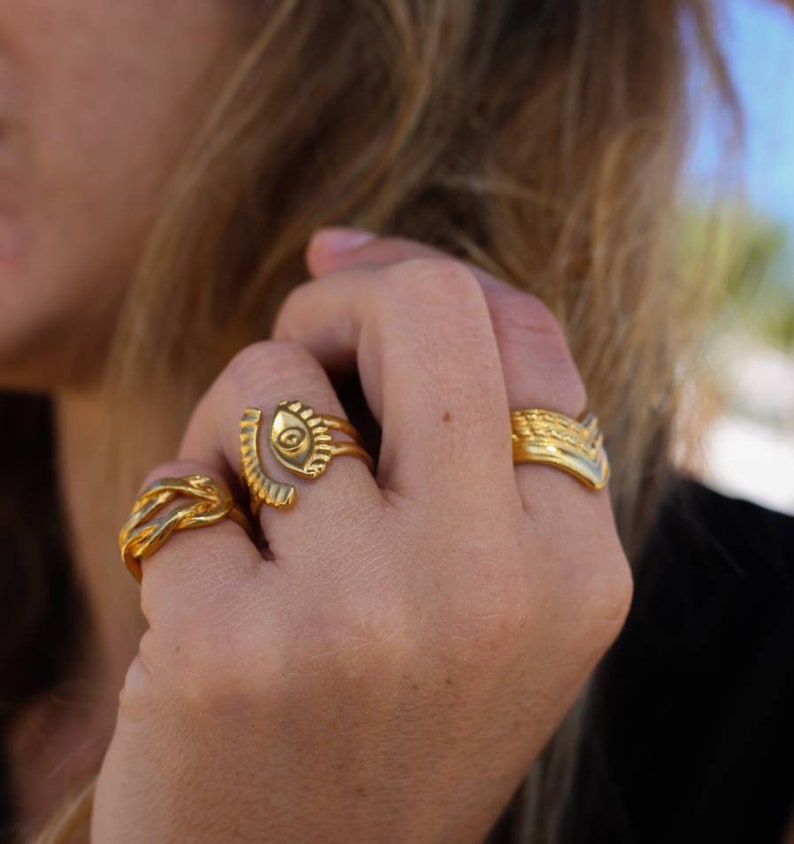 May include: A hand wearing several gold rings. The rings feature various designs, including an eye, woven patterns, and a textured band. The rings are made of a shiny gold material.