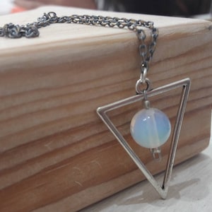 Op de afbeelding: Zilverkleurige ketting met een driehoekige hanger. Binnen de driehoek bevindt zich een ronde, opalescerende kraal. De ketting wordt gepresenteerd op een houten blok.