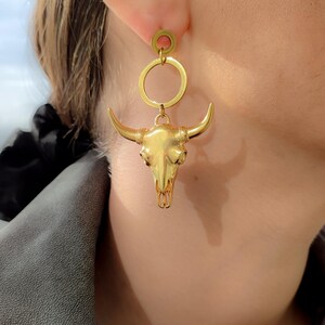 K&ouml;nnte beinhalten: Goldfarbene Ohrringe mit einem Bullensch&auml;del-Anh&auml;nger. Die Ohrringe haben eine kreisf&ouml;rmige Oberseite und ein detailliertes Bullensch&auml;del-Design. Die Ohrringe werden vor einem verschwommenen Hintergrund gezeigt.