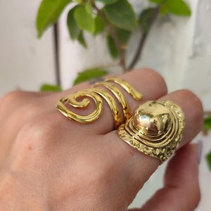 May include: Two gold-coloured rings are displayed on a hand. One ring features a spiral design, while the other has a dome-shaped top with an ornate, textured band. The rings are made of a shiny, metallic material.
