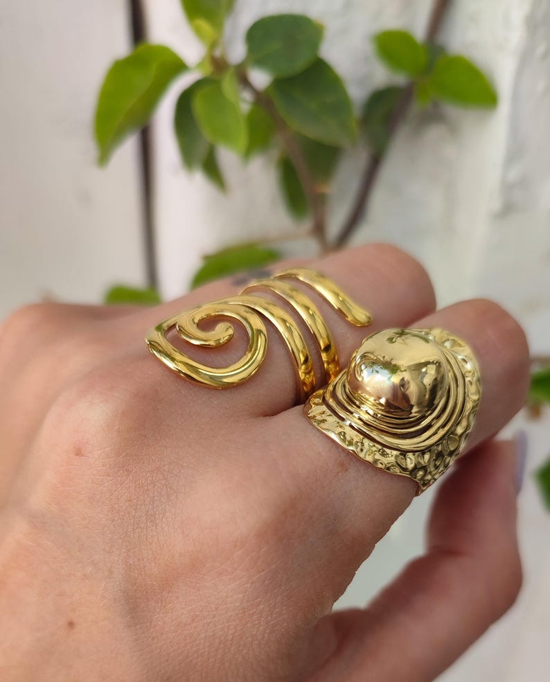 May include: Two gold-coloured rings are displayed on a hand. One ring features a spiral design, while the other has a dome-shaped top with an ornate, textured band. The rings are made of a shiny, metallic material.