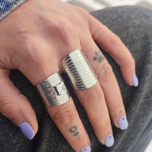 May include: Two silver rings with textured and ribbed designs. One ring features a square, clear gemstone. The rings are displayed on a hand with purple nail polish.