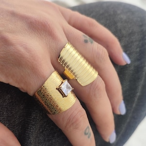 Op de afbeelding: Twee goudkleurige ringen, waarvan één met een vierkante, heldere edelsteen. Beide ringen hebben een getextureerd, geribbeld ontwerp. De ringen zitten aan een hand met lichtpaarse nagellak.