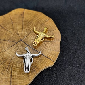 May include: Two bull skull pendants. One is gold-colored, the other is silver-colored. Both pendants have a detailed skull design with horns. The pendants are displayed on a wooden surface with a dark background.
