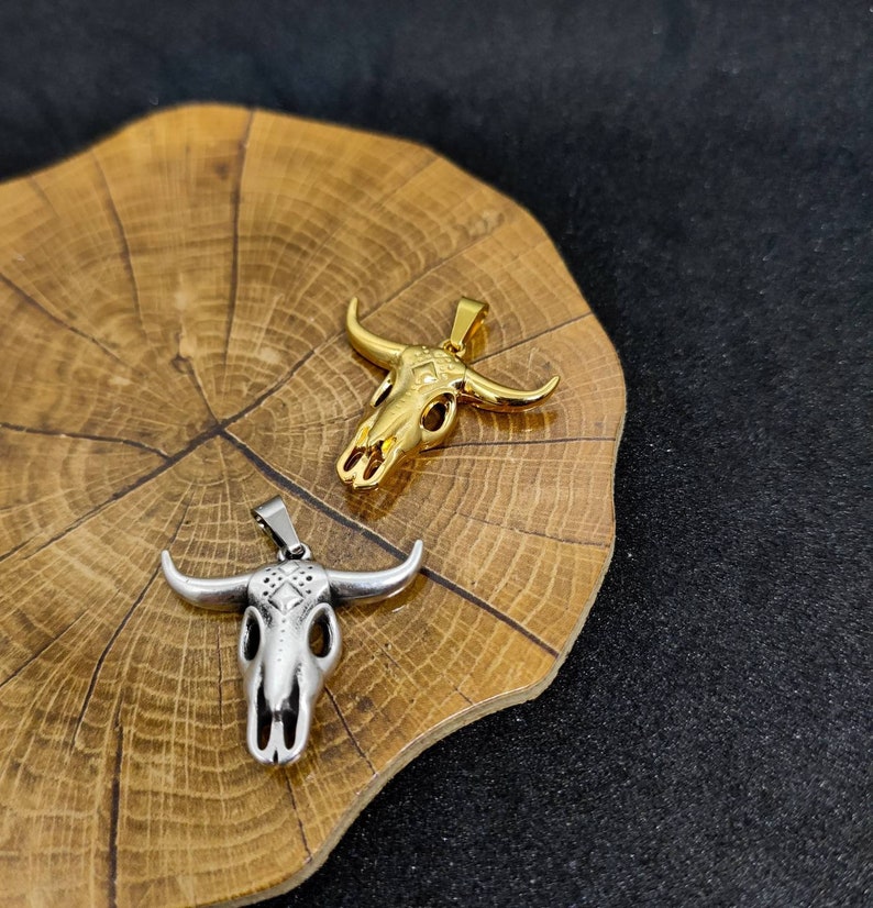May include: Two bull skull pendants. One is gold-colored, the other is silver-colored. Both pendants have a detailed skull design with horns. The pendants are displayed on a wooden surface with a dark background.