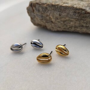 May include: Four pairs of seashell stud earrings. Two pairs are silver-toned, and two are gold-toned. The earrings are on a white surface with a gray rock in the background. The earrings are small and delicate.
