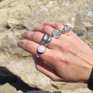 Op de afbeelding: Een hand toont verschillende zilveren ringen. E&eacute;n ring heeft een grote, ronde, lichtroze steen. Andere ringen zijn onder meer een dubbele band en twee zegelringen met reli&euml;fpatronen. De achtergrond toont een rotsachtig oppervlak en water.