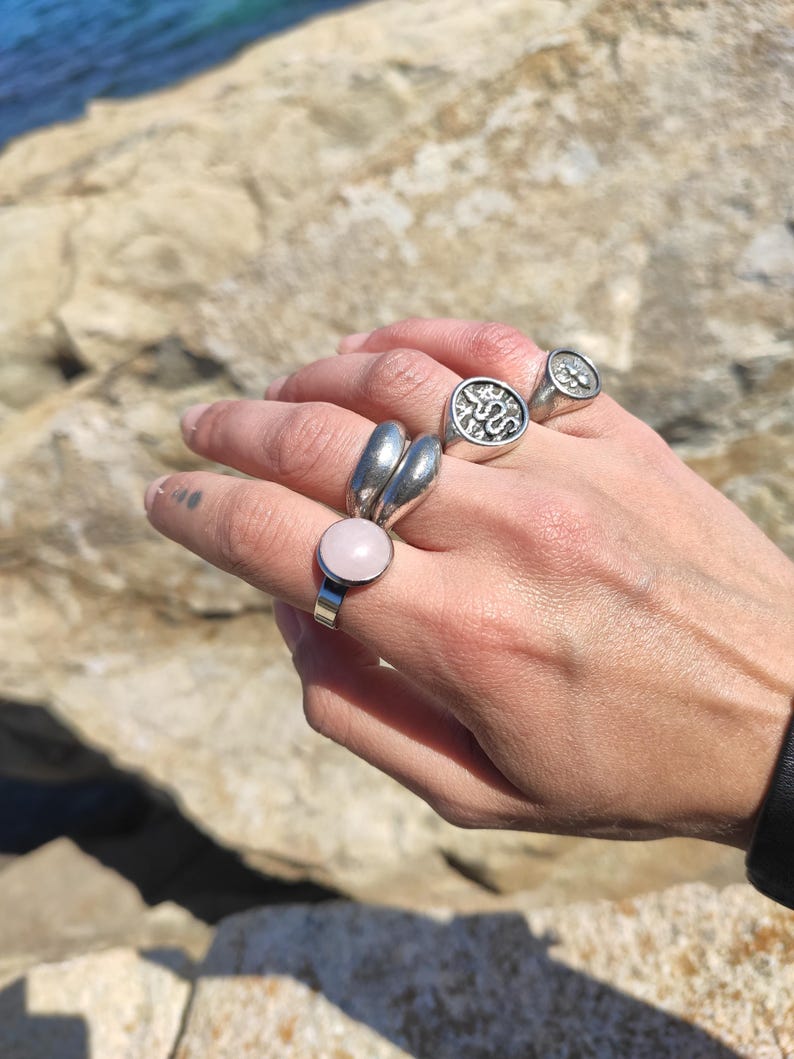Op de afbeelding: Een hand toont verschillende zilveren ringen. E&eacute;n ring heeft een grote, ronde, lichtroze steen. Andere ringen zijn onder meer een dubbele band en twee zegelringen met reli&euml;fpatronen. De achtergrond toont een rotsachtig oppervlak en water.