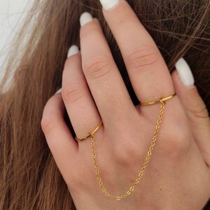 May include: A gold double-finger ring featuring a fine chain linking two bands. The ring is displayed on a hand with white nail polish, with brown hair in the background. The jewellery item is simple and elegant.