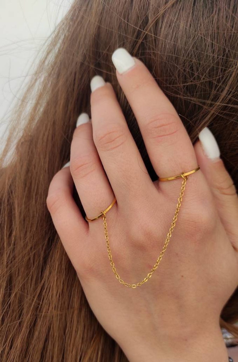 May include: A gold double-finger ring featuring a fine chain linking two bands. The ring is displayed on a hand with white nail polish, with brown hair in the background. The jewellery item is simple and elegant.