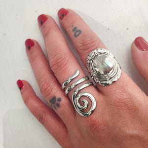 May include: Two silver rings on a hand. One ring is a spiral design and the other is a round ring with a raised center.