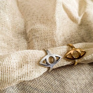 May include: Two rings with an eye design, one silver and one gold, rest on a beige, textured fabric. The rings have a stylized eye shape with a circular center and a curved band.