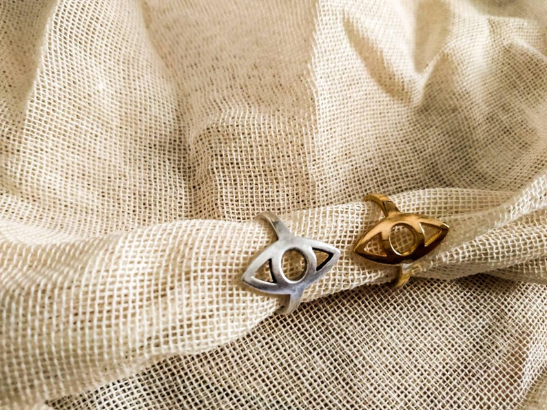 May include: Two rings with an eye design, one silver and one gold, rest on a beige, textured fabric. The rings have a stylized eye shape with a circular center and a curved band.