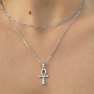 Könnte beinhalten: Ein silbernes Halsketten-Set mit zwei Ketten. Die obere Kette hat ein Design mit Büroklammer-Gliedern, während die untere Kette einen Anhänger in Form eines Ankh-Kreuzes trägt. Der Schmuck wird vor einem Hautton-Hintergrund präsentiert.
