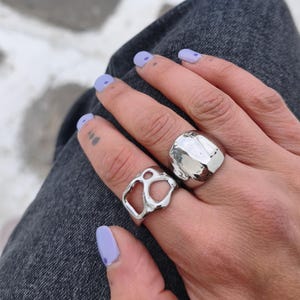 May include: Two silver rings are shown on a hand. One ring features an organic, open design with irregular shapes. The other ring is a large, solid, rounded shape with a textured surface. The hand has light purple nail polish.