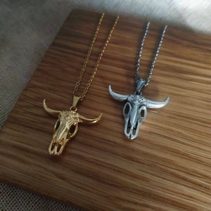 May include: Two necklaces with bull skull pendants. One is gold-colored, the other is silver-colored. Both pendants have detailed skull features and are suspended from delicate chains. The necklaces are displayed on a wooden surface.