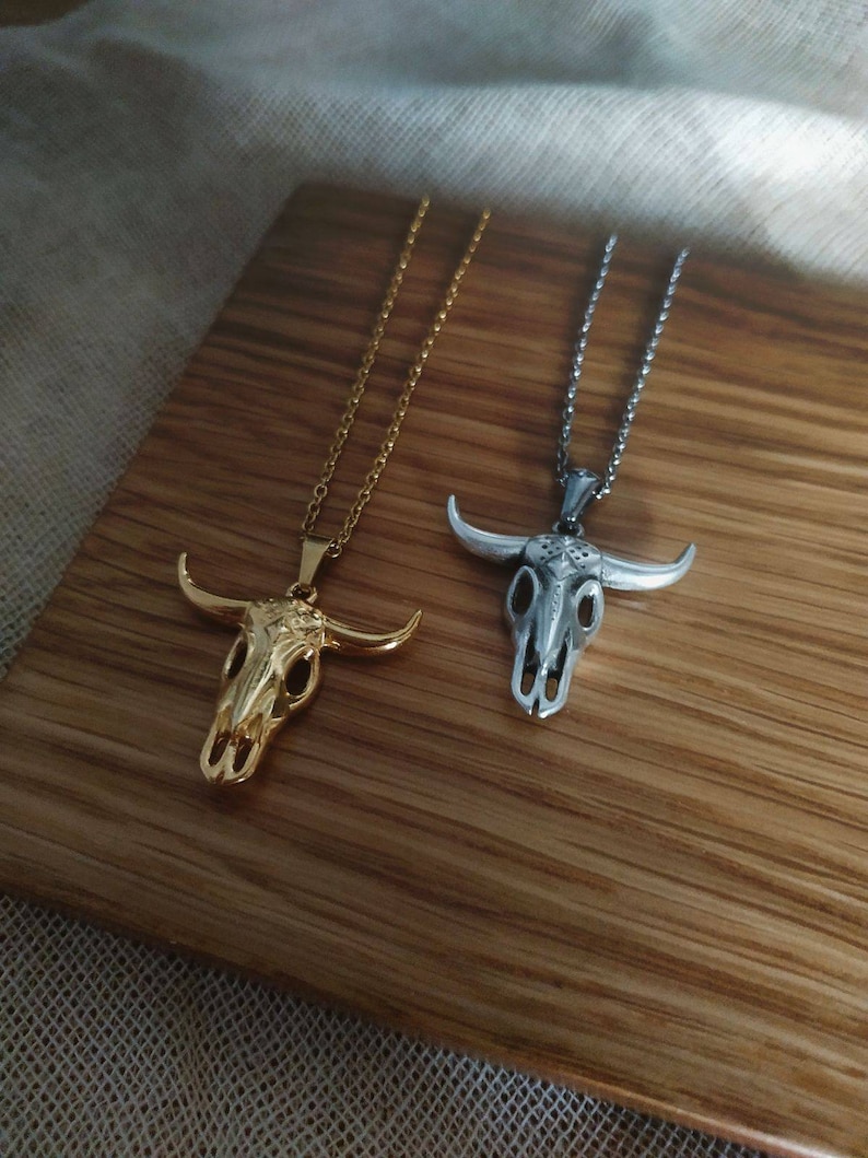 May include: Two necklaces with bull skull pendants. One is gold-colored, the other is silver-colored. Both pendants have detailed skull features and are suspended from delicate chains. The necklaces are displayed on a wooden surface.