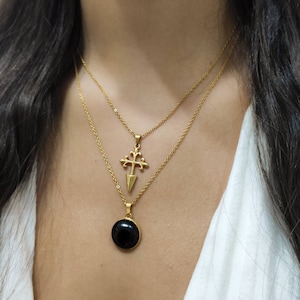 May include: Two gold-colored necklaces layered together. The top necklace features a pendant with a cross design. The bottom necklace has a round, black stone pendant. Both necklaces are on a person wearing a white top.