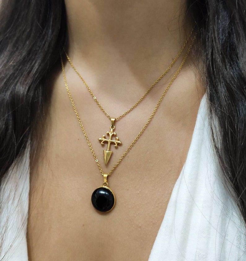 May include: Two gold-colored necklaces layered together. The top necklace features a pendant with a cross design. The bottom necklace has a round, black stone pendant. Both necklaces are on a person wearing a white top.