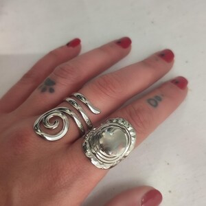 May include: Two silver rings on a finger. One ring is a spiral design and the other is a round ring with a textured surface.